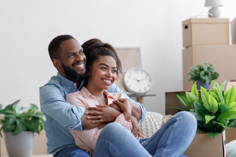 Satisfied young black husband and wife hugging sit on floor with cardboard boxes things and plants i